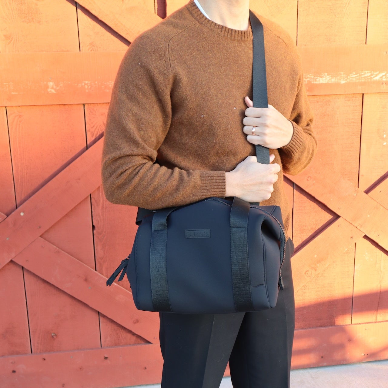 Person wearing a brown sweater and dark pants holding a black duffle diaper bag in front of a wooden door.