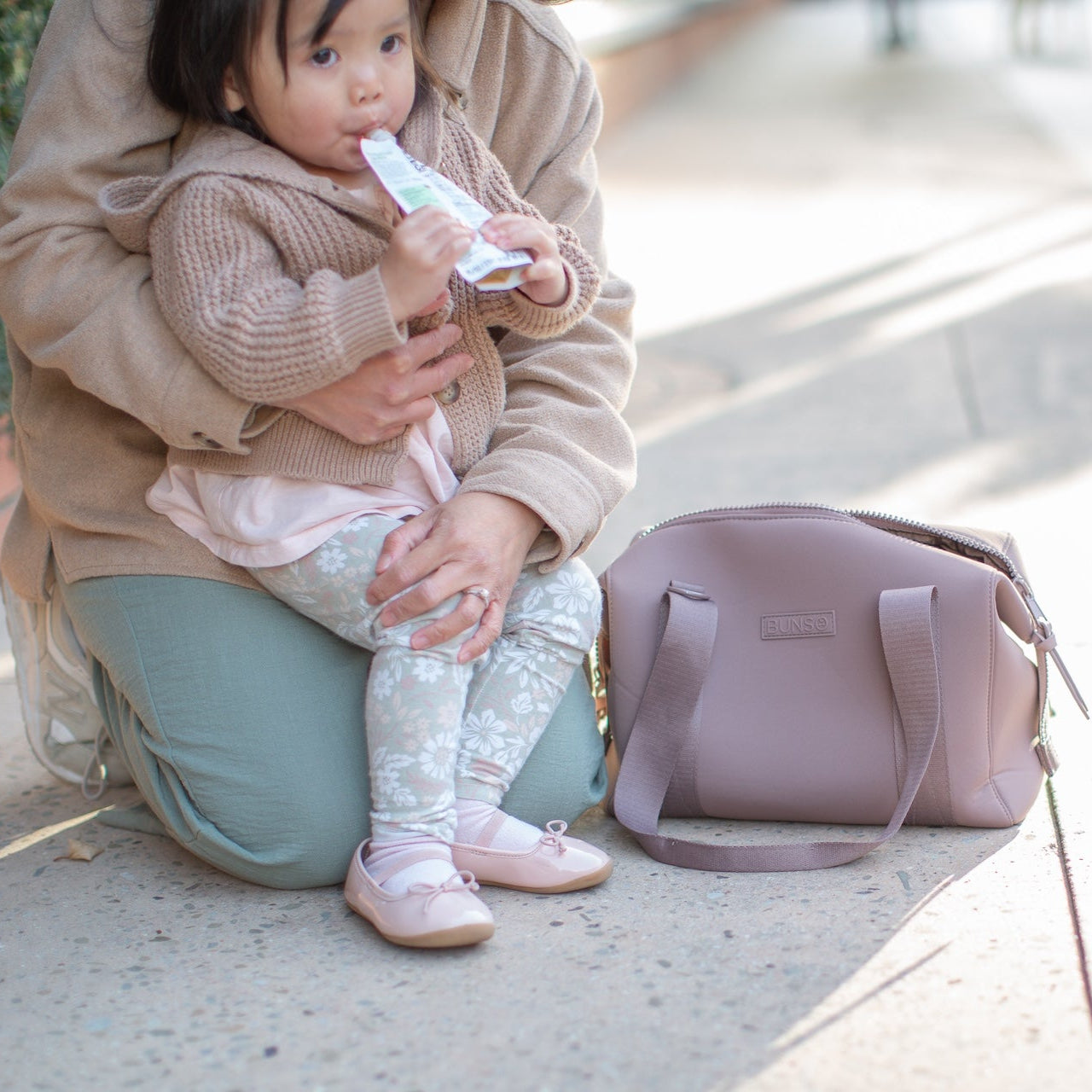 A woman holding a child next to a mauve duffel bag on a sidewalk.