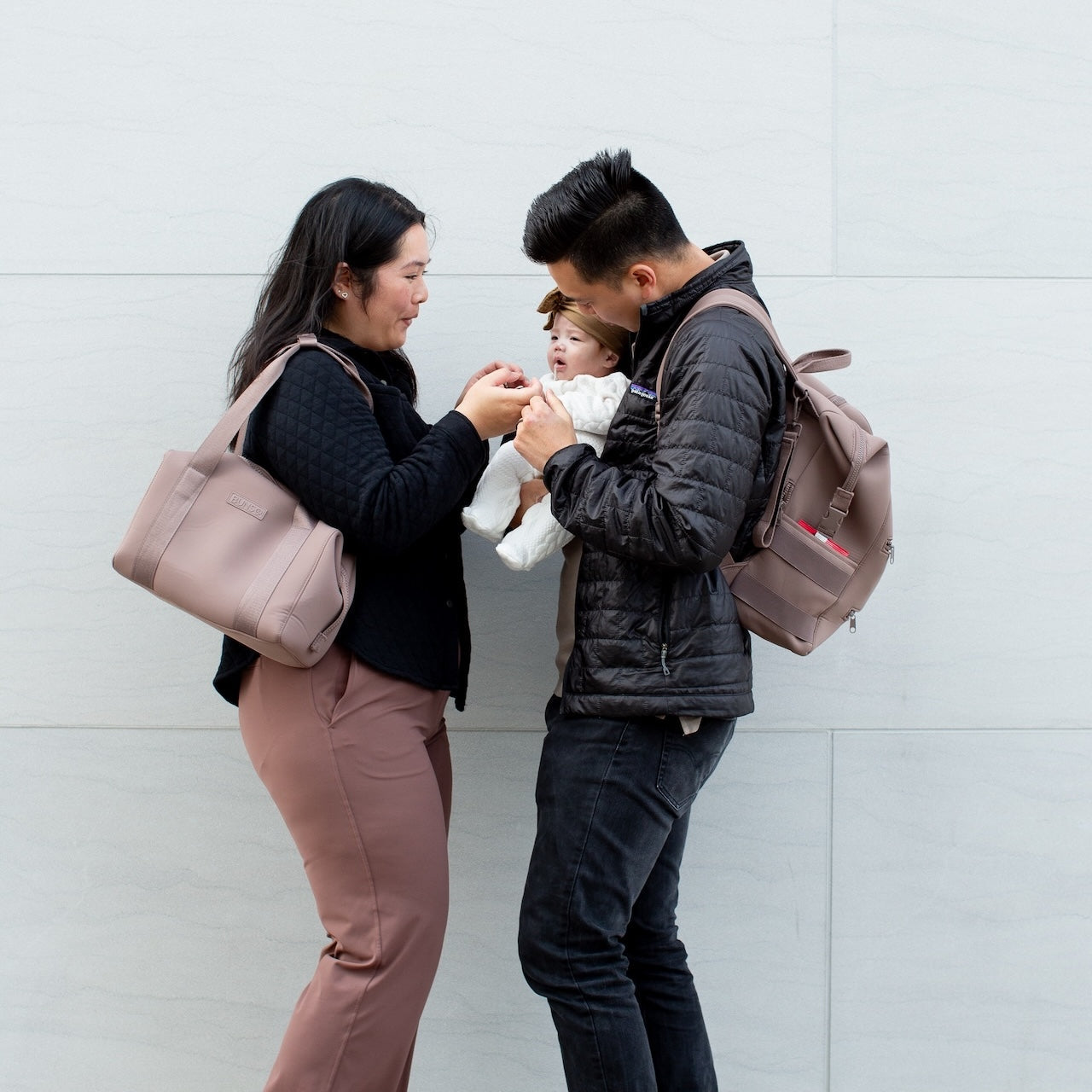 Parents are carrying their child while standing together against a white wall.