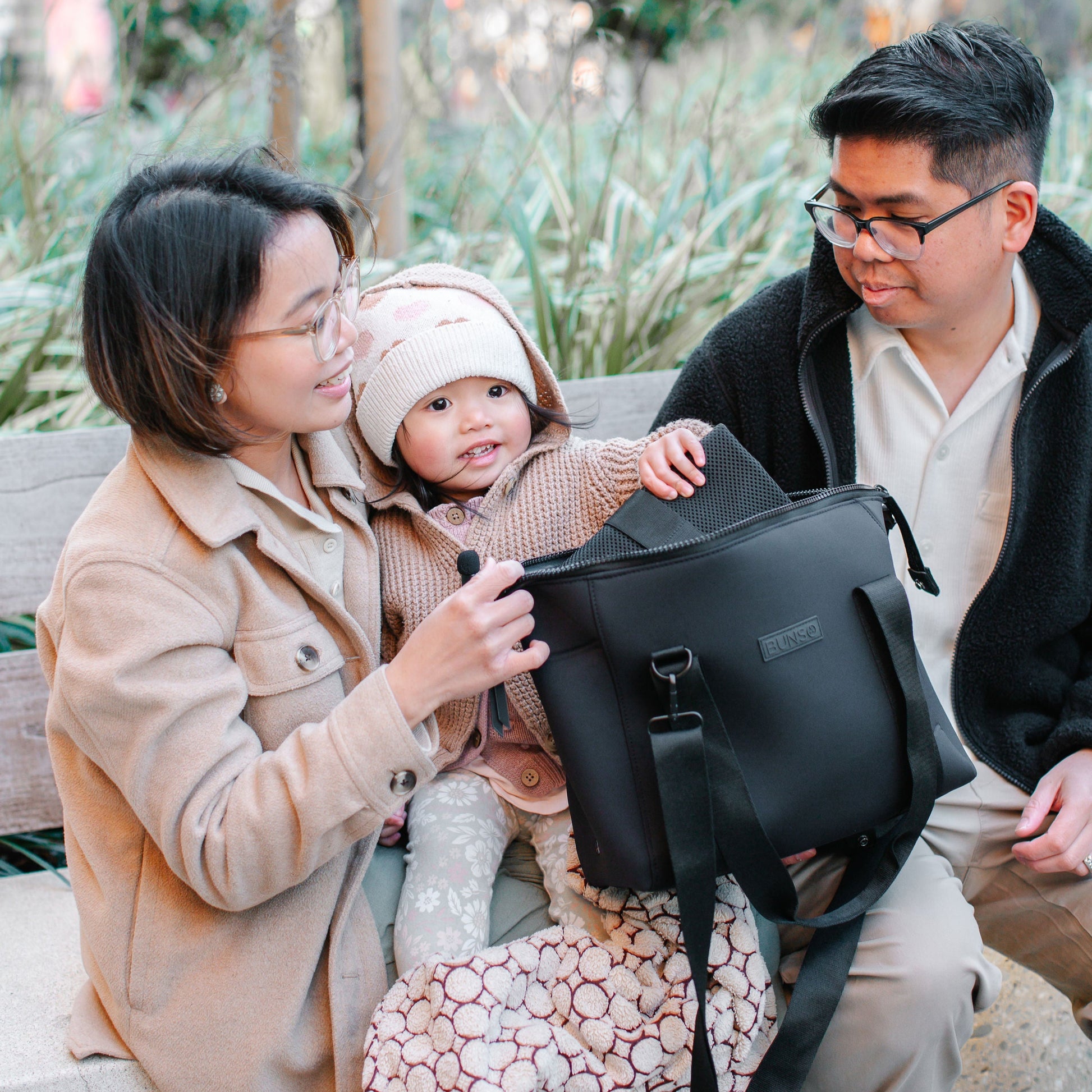 A family of three with a black duffel diaper bag in an outdoor setting.