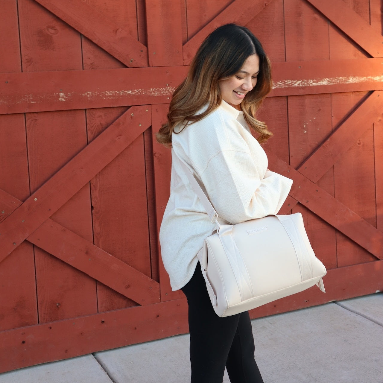 A woman wearing a cream duffel diaper bag in front of a red wooden door.