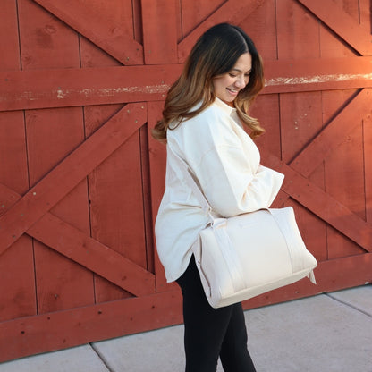 A woman wearing a cream duffel diaper bag in front of a red wooden door.